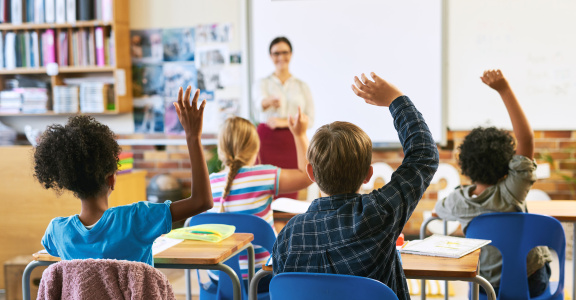 A teach in the front of a classroom with students raising their hands